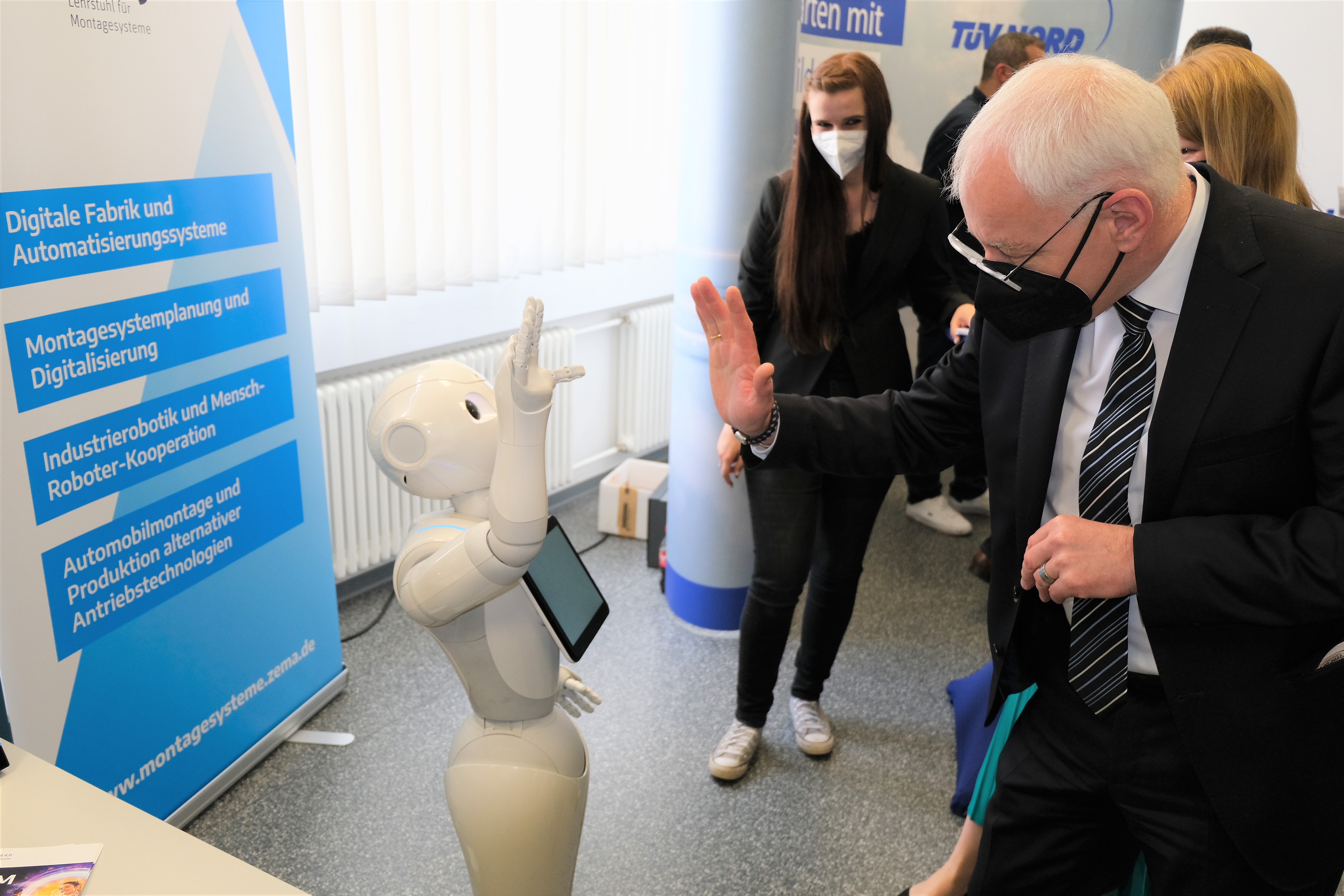 Mann mit Maske hebt Hand und gegenüber hebt ein Roboter auch die Hand zum Gruße